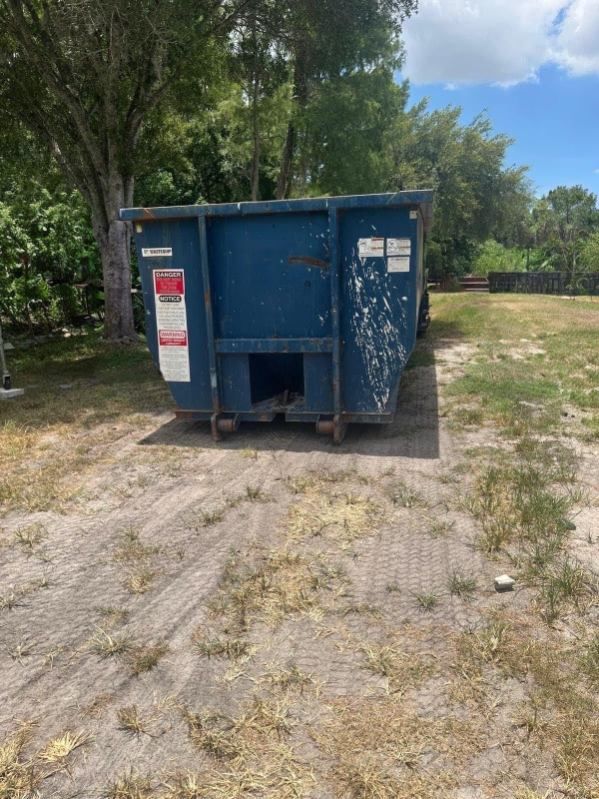 Blue dumpster on a gravel lot beside trees under a partly cloudy sky