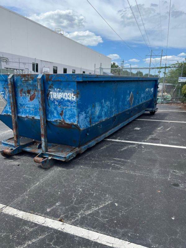 Blue industrial dumpster in a parking lot beside a white building under a cloudy sky
