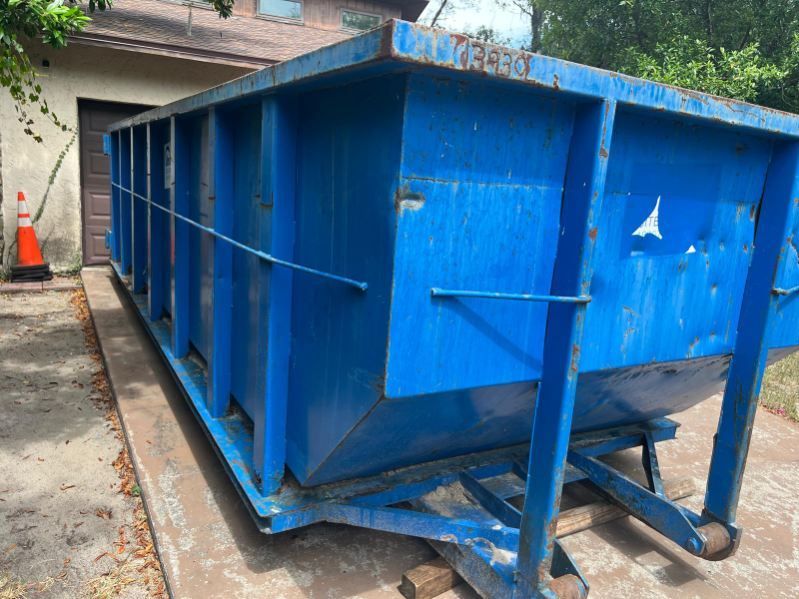 Blue industrial dumpster on a concrete lot beside a building, with a traffic cone in the background.