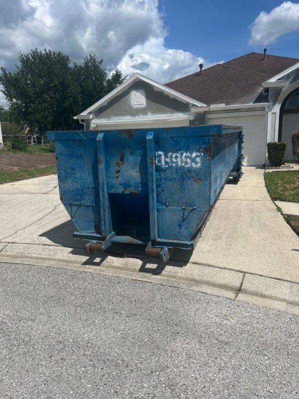 Blue dumpster on a driveway in front of a suburban house under a partly cloudy sky