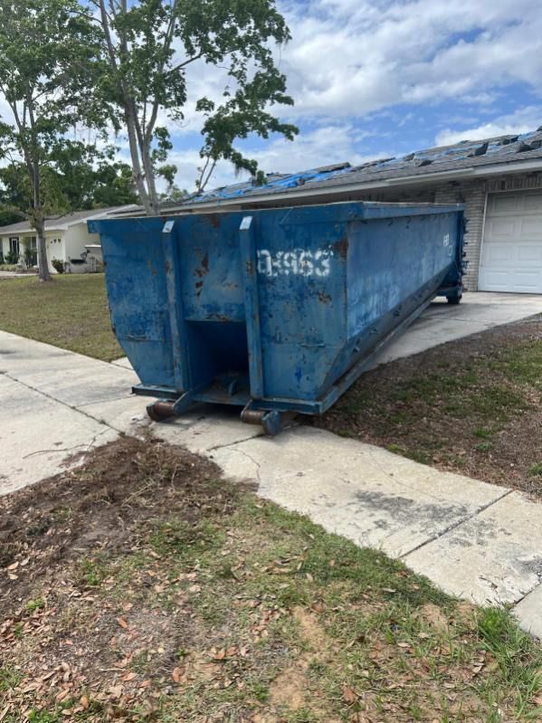 Blue dumpster on a sidewalk beside a house and driveway, with the number 49953 on the front.