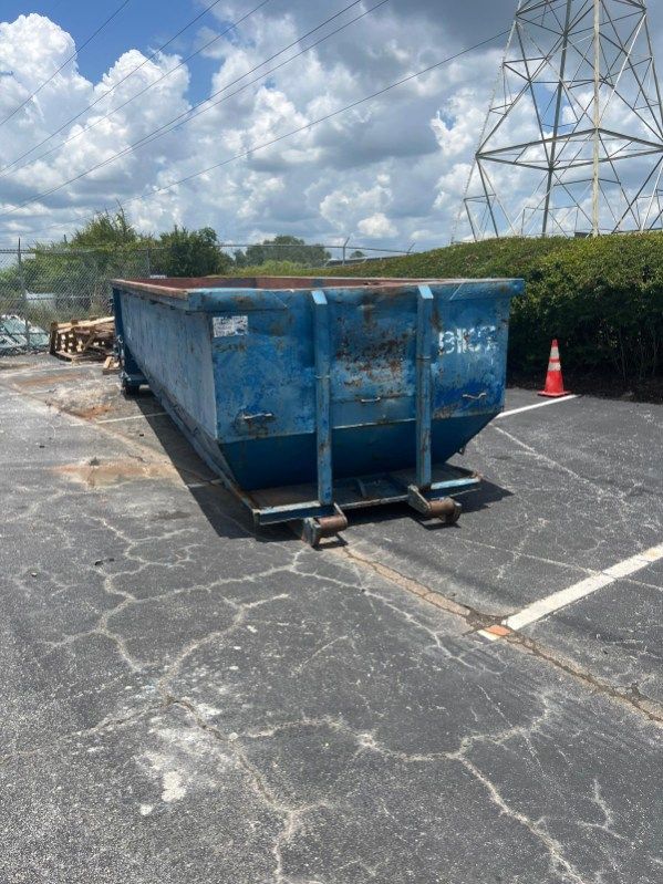 Blue dumpster in a parking lot with cracked asphalt, a traffic cone, and power lines in the background
