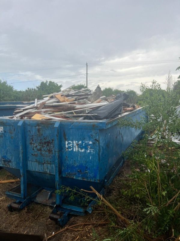 Blue dumpster filled with debris and scrap metal in an overgrown outdoor lot under a cloudy sky