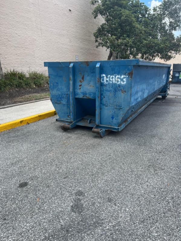 Large blue metal dumpster on an asphalt lot beside a beige building and yellow curb.