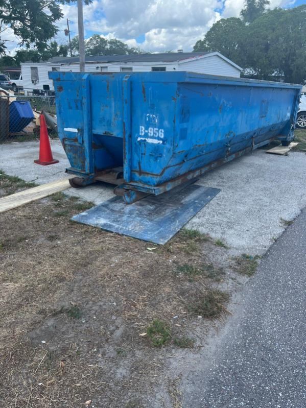Blue construction dumpster on a concrete pad beside a road, with orange cones and a house in the background