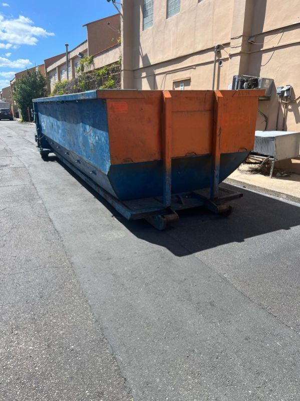 Large blue and orange industrial dumpster on a sunlit street beside beige buildings