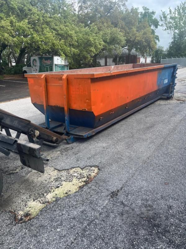Orange-and-blue dumpster on a cracked road beside trees, with a damaged front loader in the foreground.