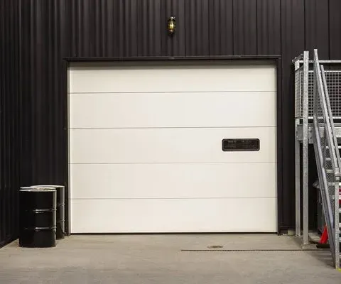 White garage door on a black building with a metal staircase to the right and barrels on the left.