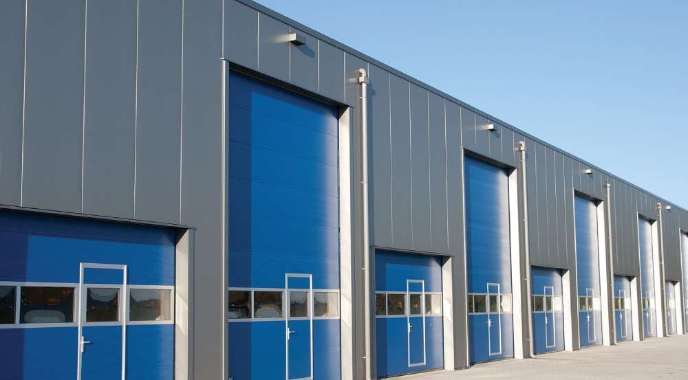 Row of industrial buildings with blue overhead doors and gray metal siding under a clear blue sky.