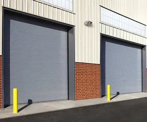 Two gray warehouse roll-up doors, each flanked by a yellow bollard, on a brick and metal building.