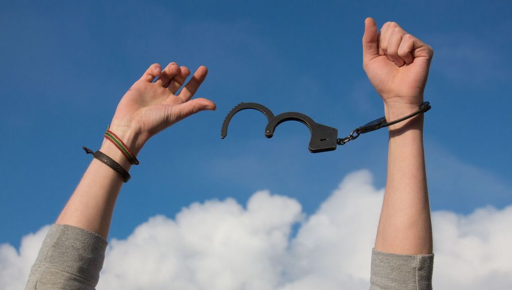 Hands in handcuffs raised against a blue sky with clouds