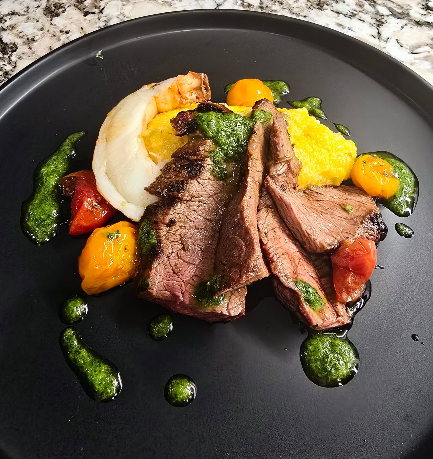 Grilled steak with shrimp, polenta, tomatoes, and green sauce on a black plate.