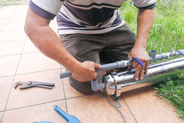 Technician installing stainless steel water pump with PVC pipes and valve in outdoor plumbing setup.