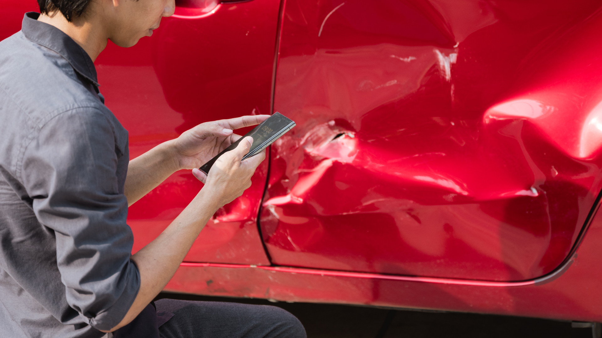 Man examining damage on a red car door, holding a phone.