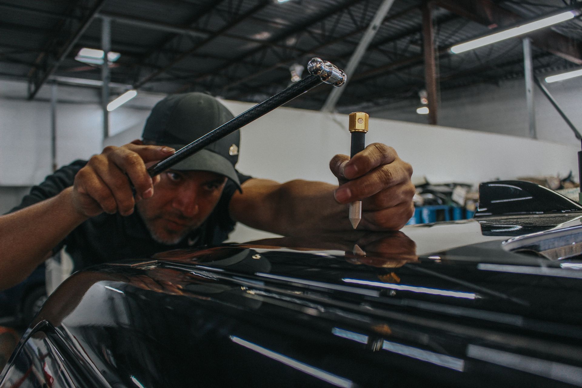 Mechanic polishing a black car hood in a garage, using a handheld tool.