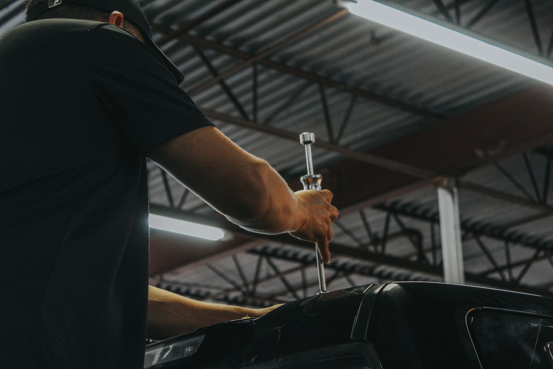 Person using a spray bottle in a garage, with a car hood in the foreground and roof beams overhead