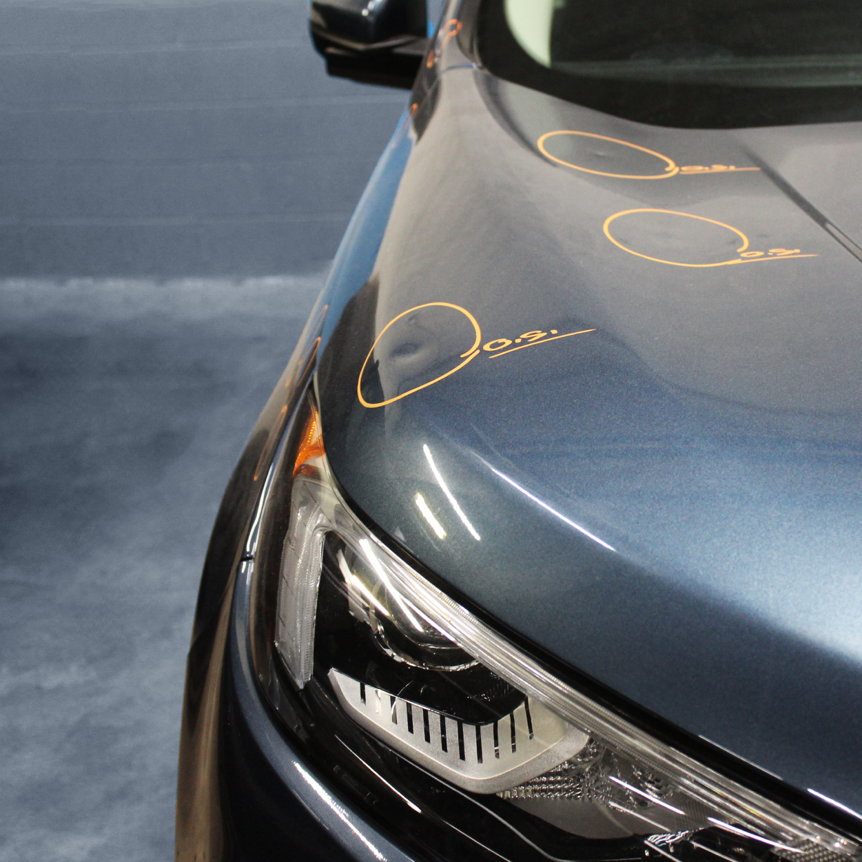 Close-up of a blue car hood and headlight with orange marker circles on a showroom floor