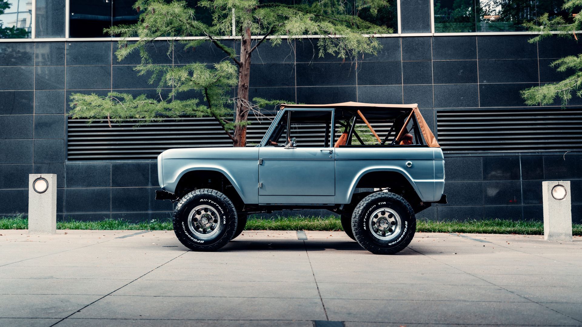 A blue vintage Ford Bronco SUV parked on a city street in front of a modern building.