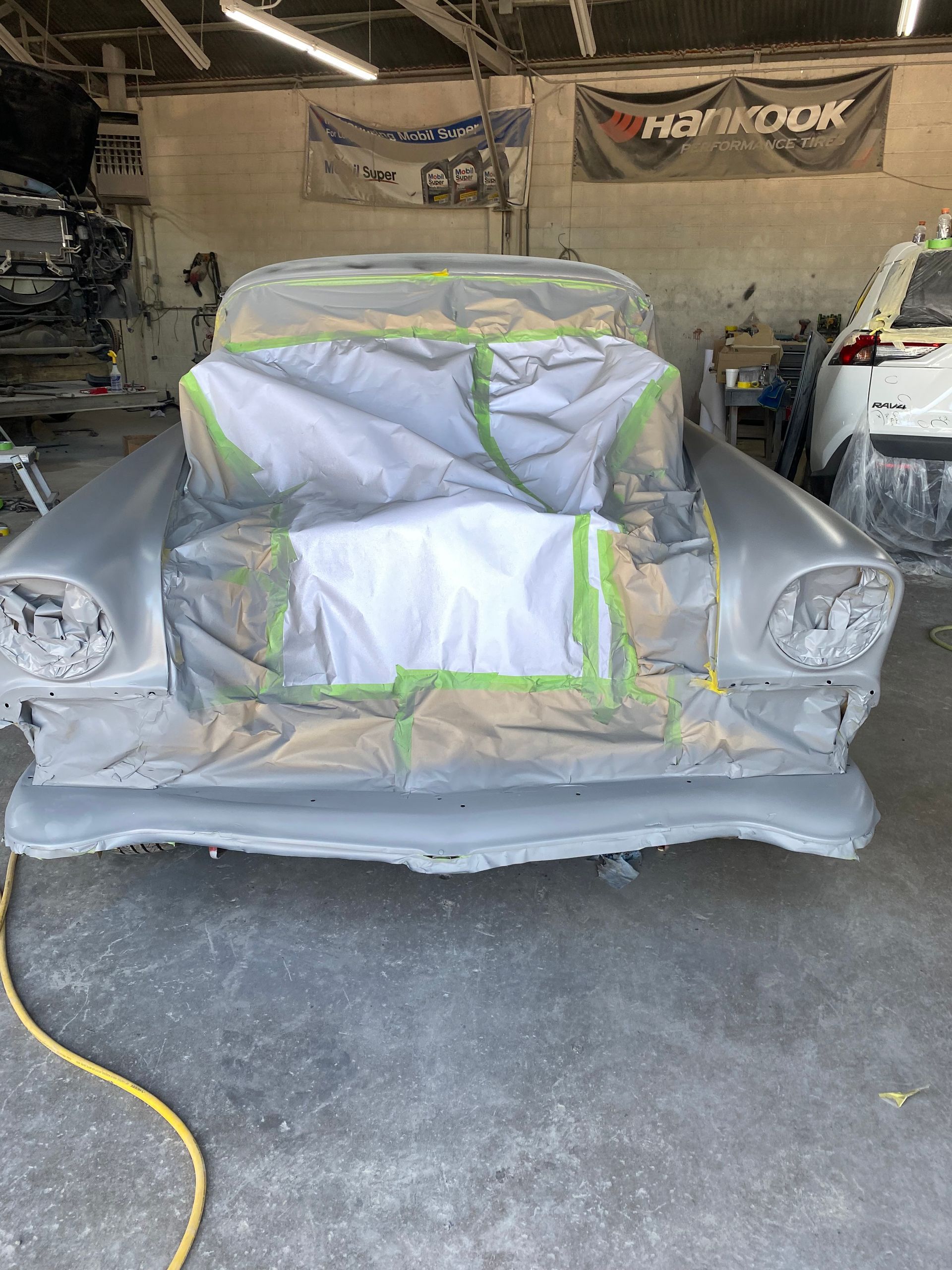 A classic car prepped for paint, masked with paper and tape, in a garage.