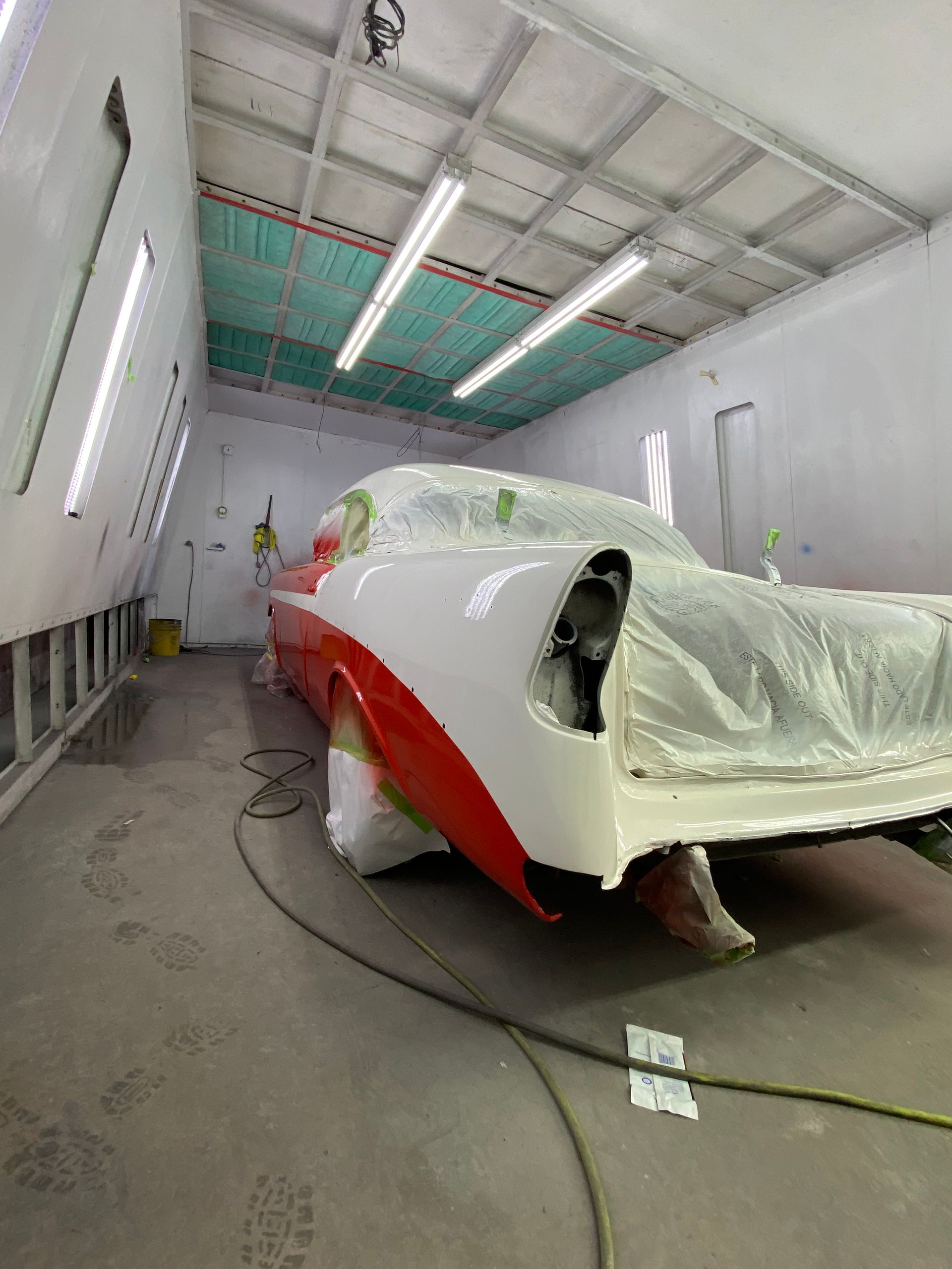 Classic car, red and white, in a spray paint booth. Bodywork is taped and covered, ready for paint.
