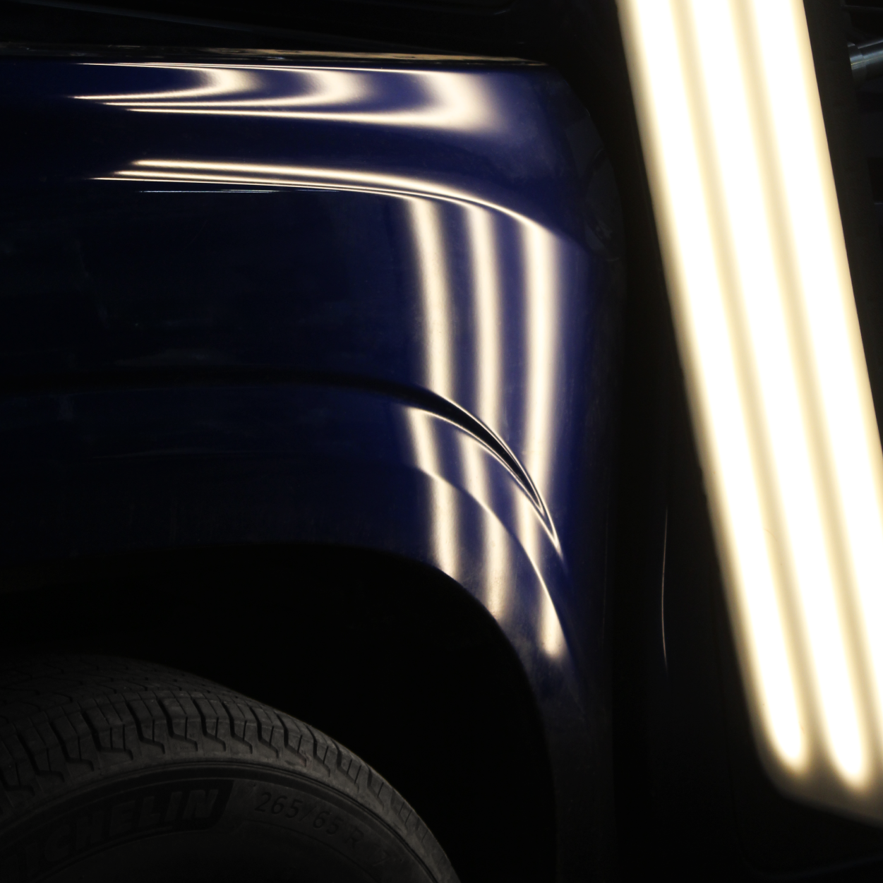 Close-up of a dark blue car fender with bright vertical light reflections and part of a tire visible