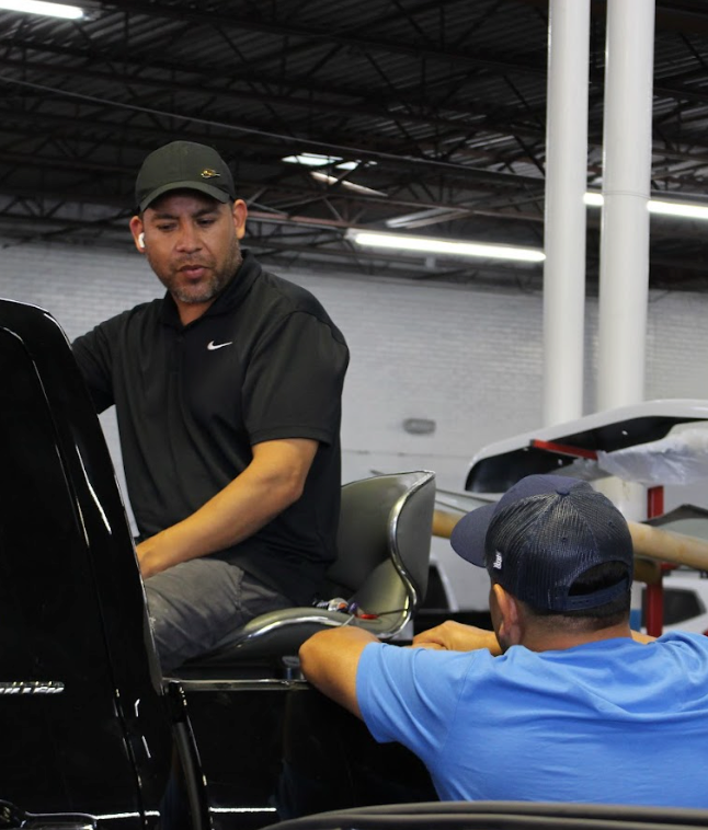 Two men in a garage, one wearing black shirt looking at car, other wearing blue shirt, both focused.