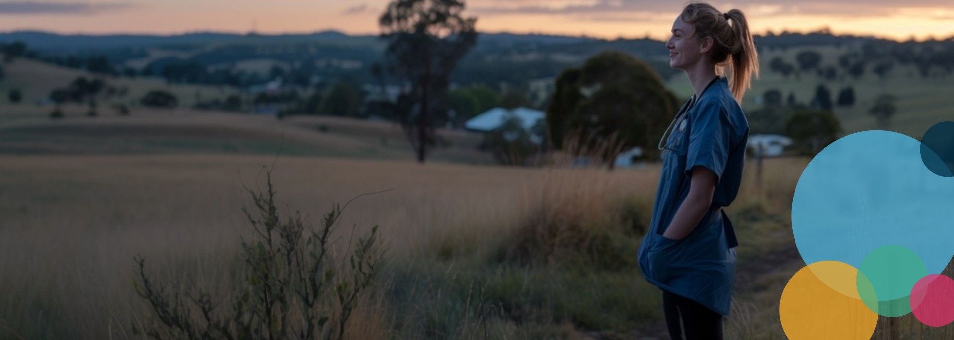 Female nurse stands on a grassy hill, hands in pockets, looking at a rural landscape at dusk.