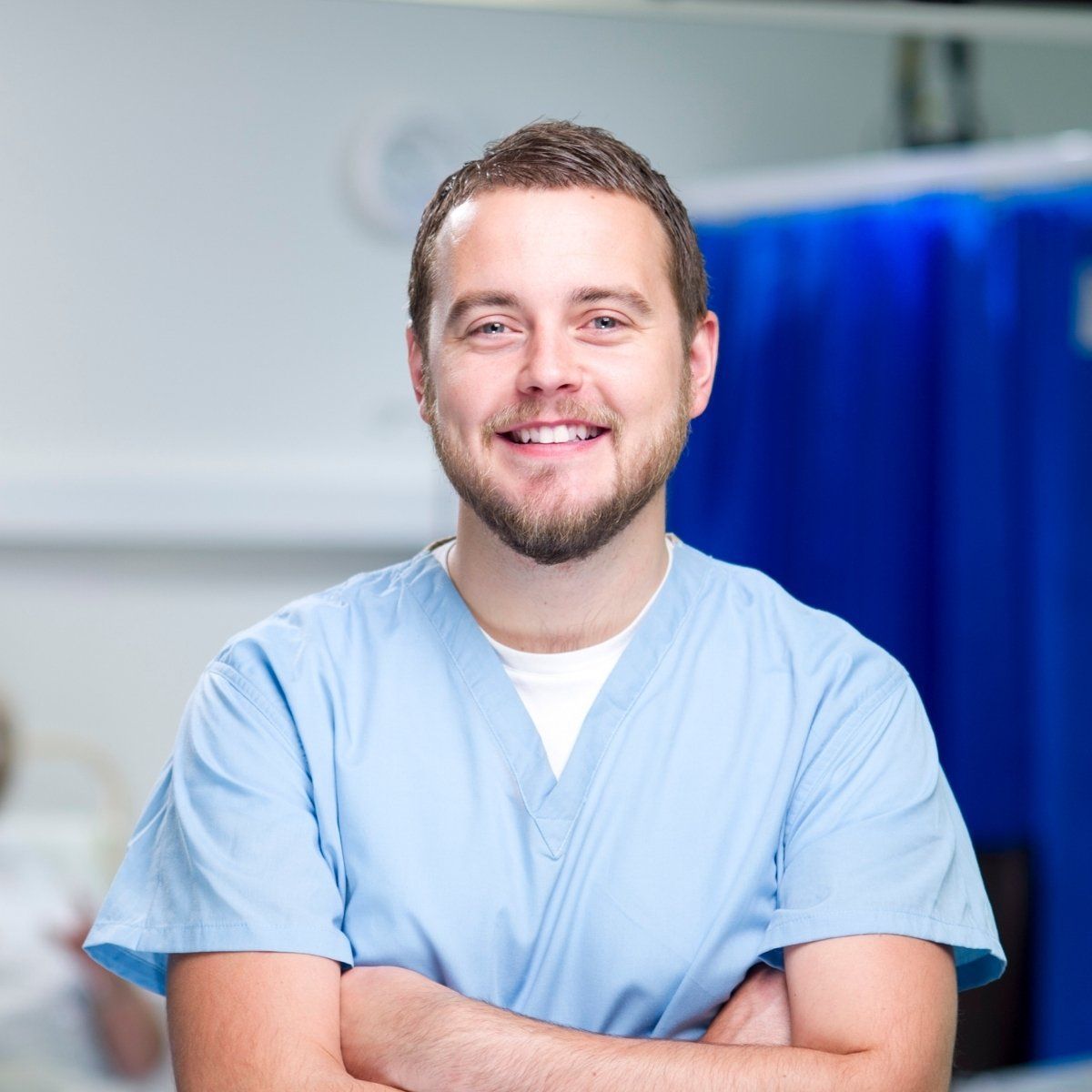 Smiling healthcare worker in blue scrubs with arms crossed, in a medical setting.