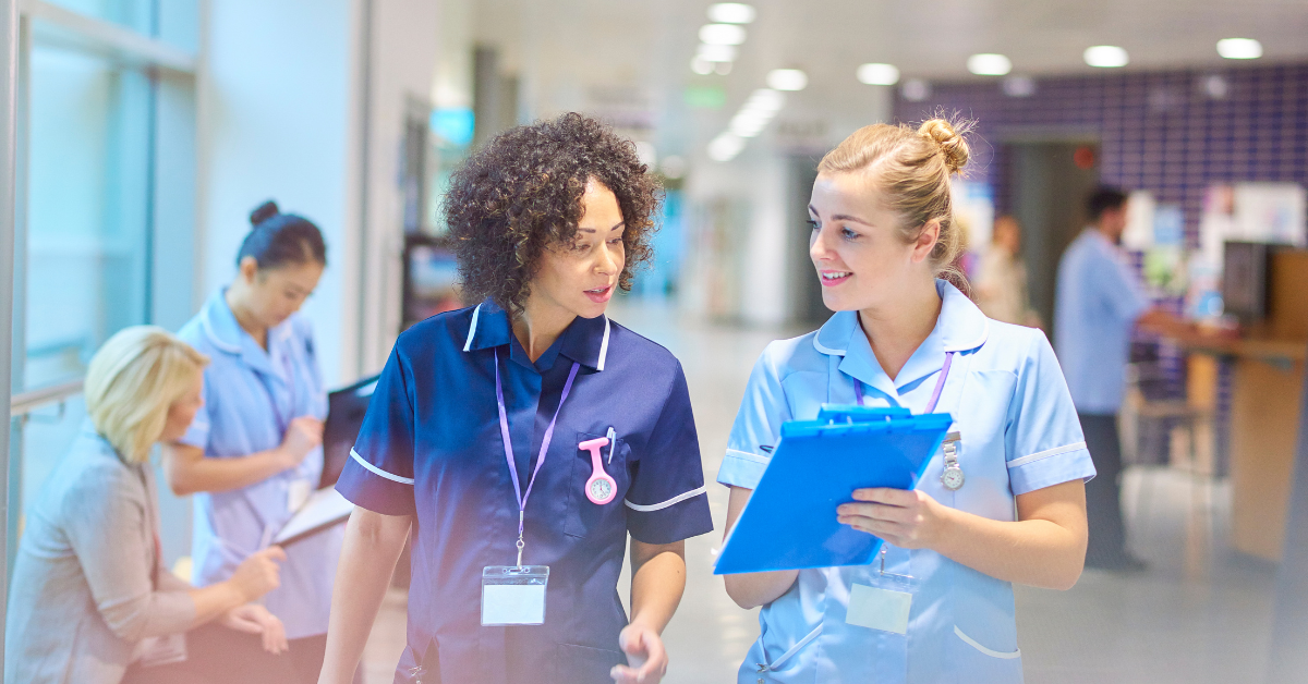 Two healthcare workers in scrubs walking and talking in a hospital hallway, one holding a blue clipboard.