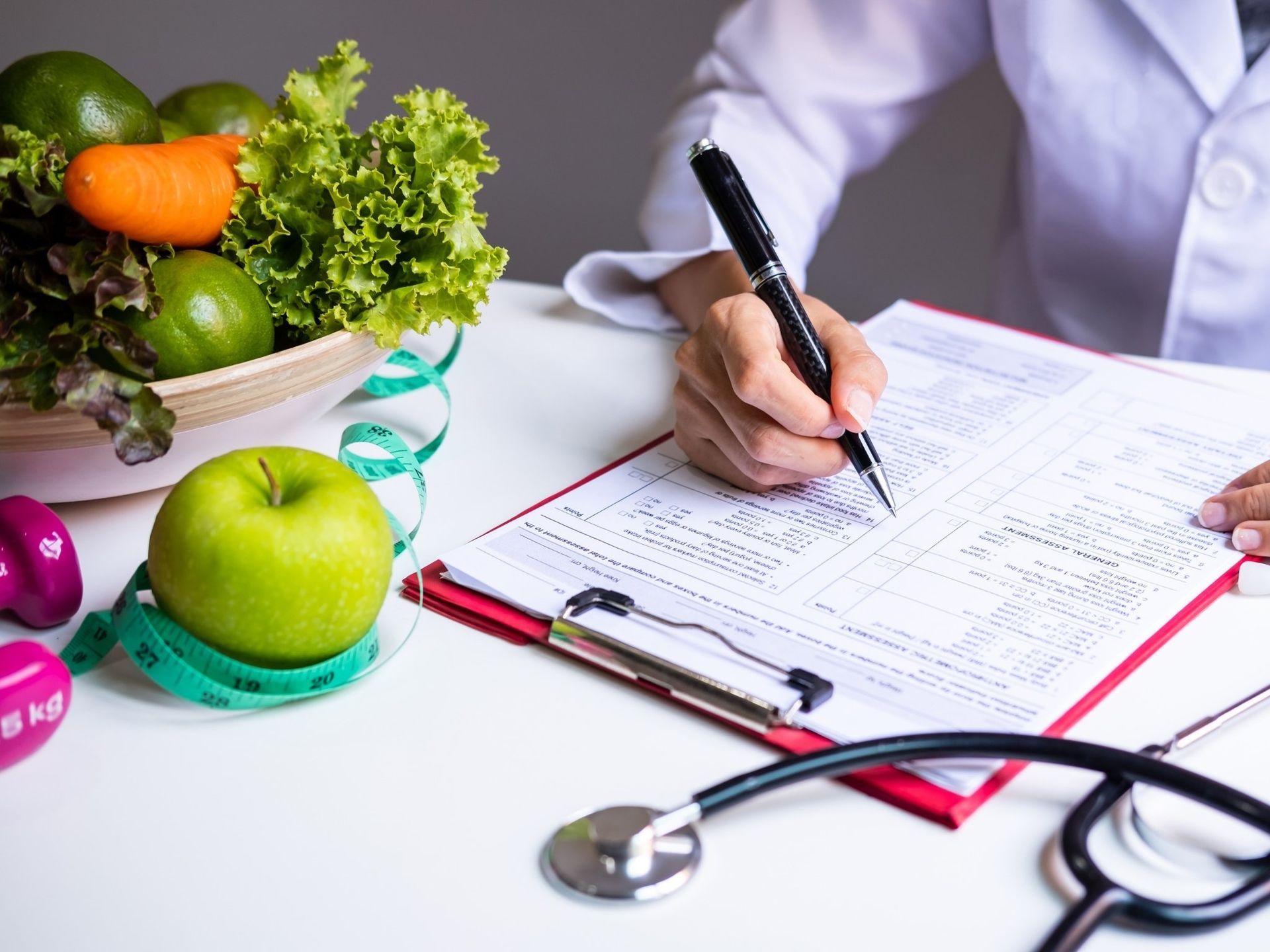 A doctor is writing on a clipboard next to a stethoscope, fruits and weights.