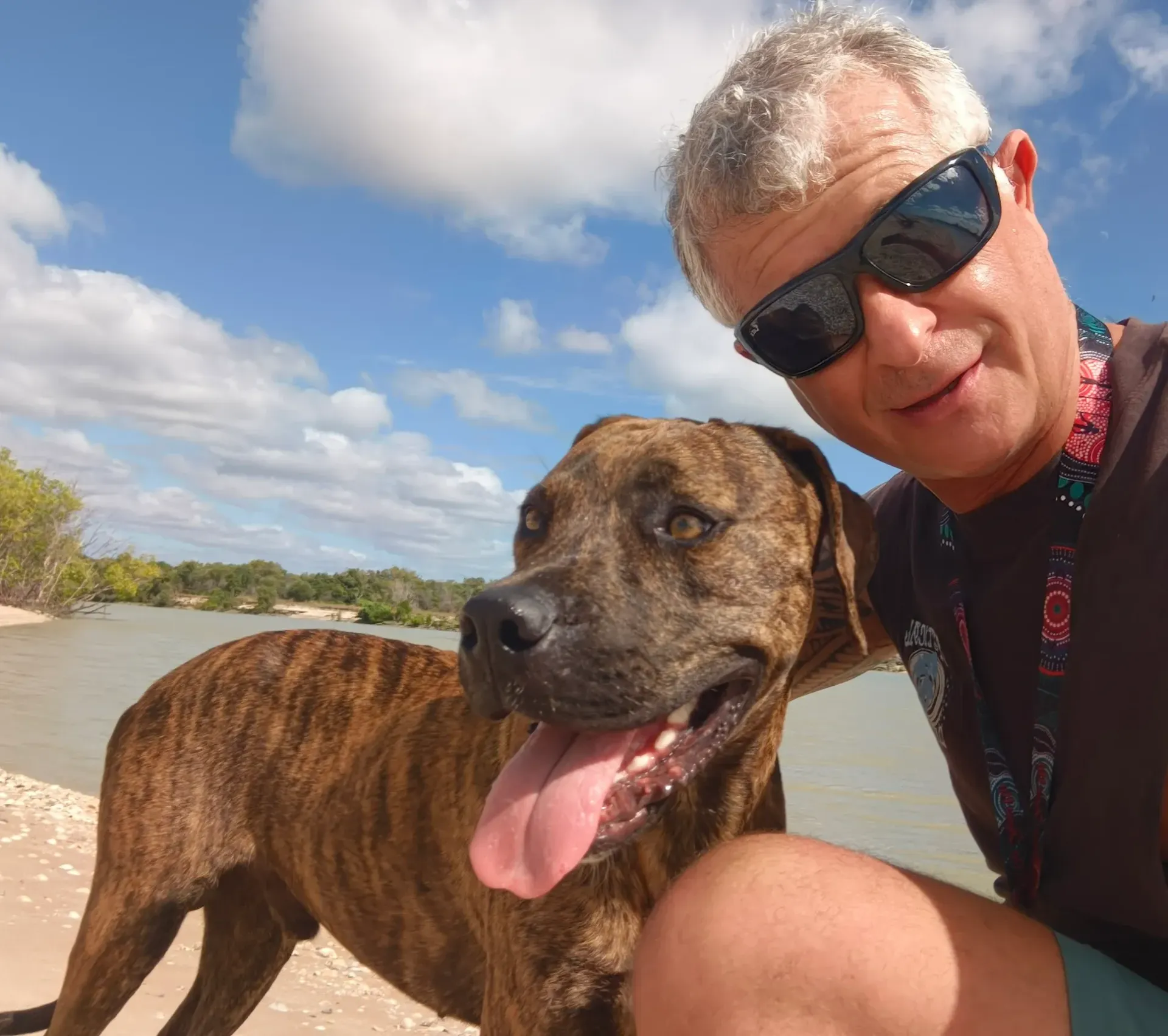 A man wearing sunglasses poses for a selfie with a brindle dog with its tongue out on a sunny beach near water.
