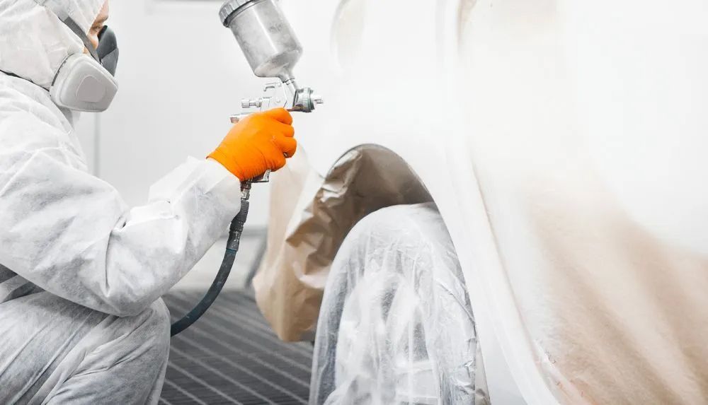 Person in protective suit spray-painting a car's covered tire; orange gloves, white setting.