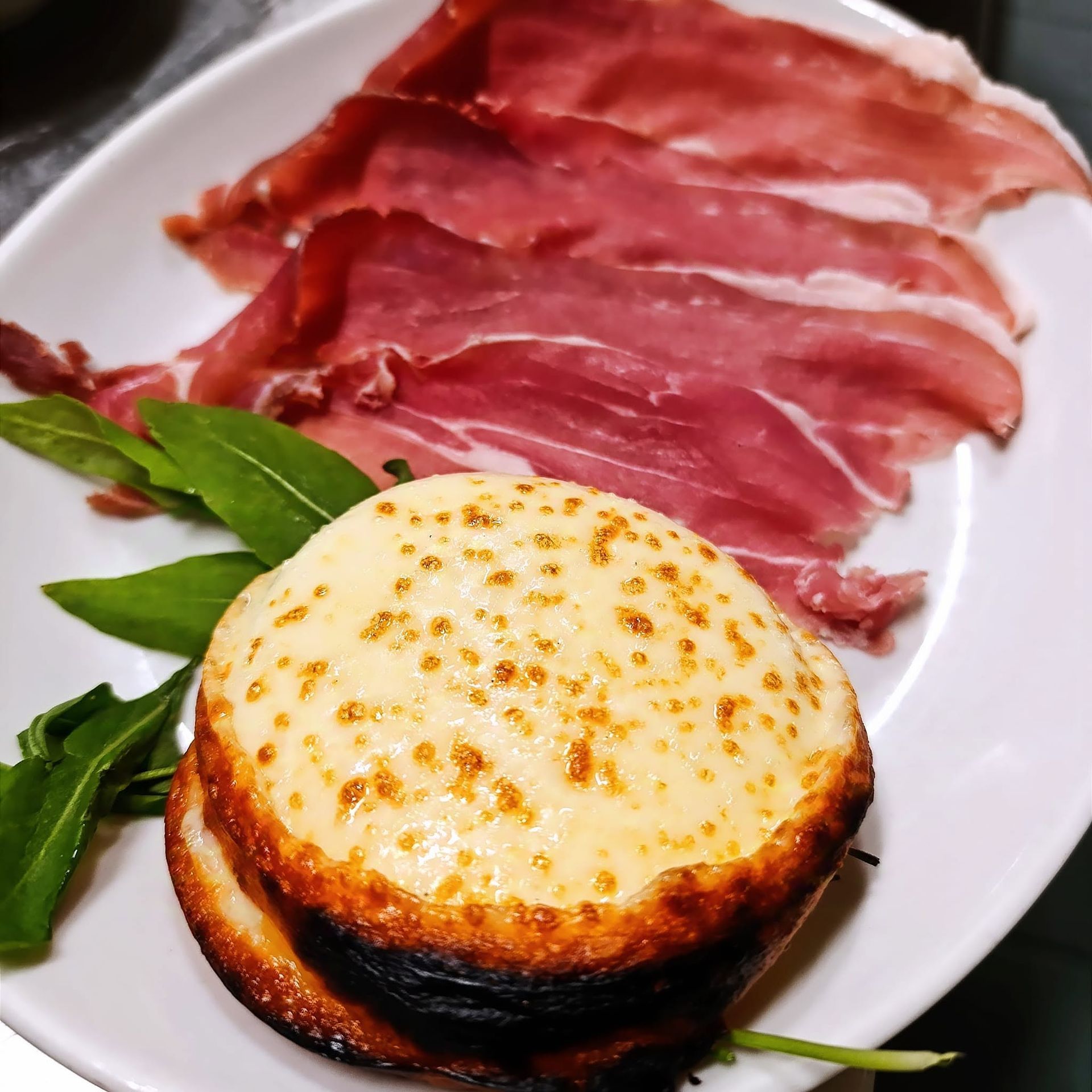 Antipasto rotondo con formaggio scottato, affettati e affettati, servito su un piatto bianco e guarnito con foglie verdi.