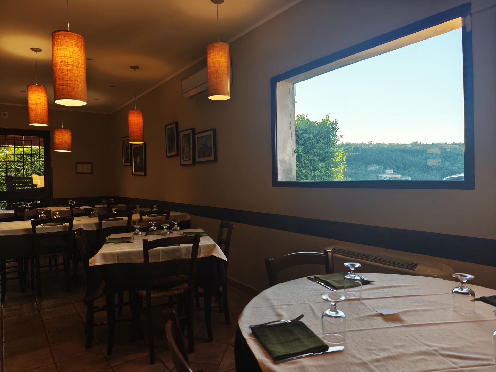 Sala da pranzo vuota di un ristorante, con tavoli e calde lampade a sospensione, affacciata su una luminosa finestra.