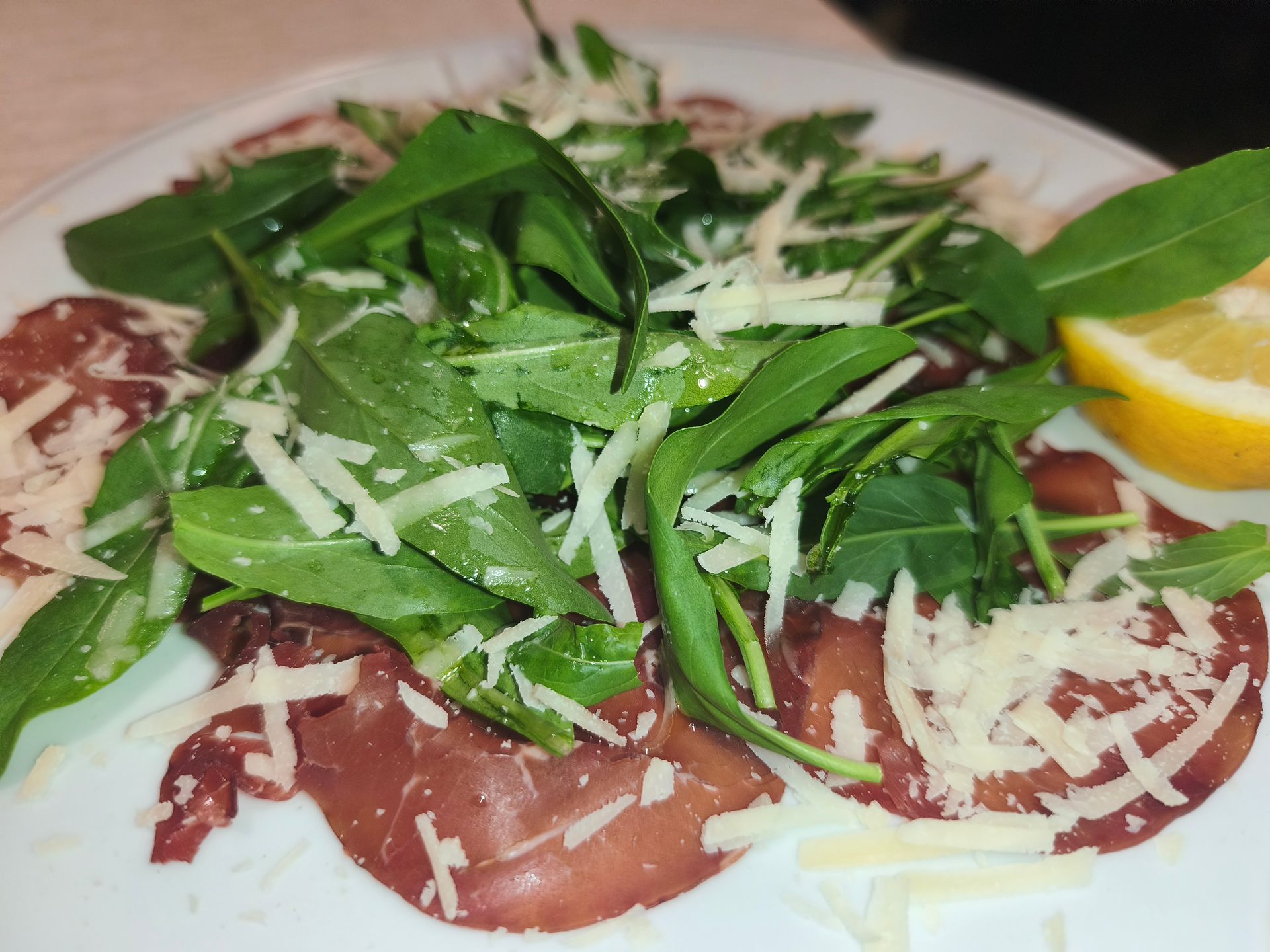 Carpaccio di manzo tagliato finemente con rucola, scaglie di formaggio e spicchi di limone su un piatto bianco.