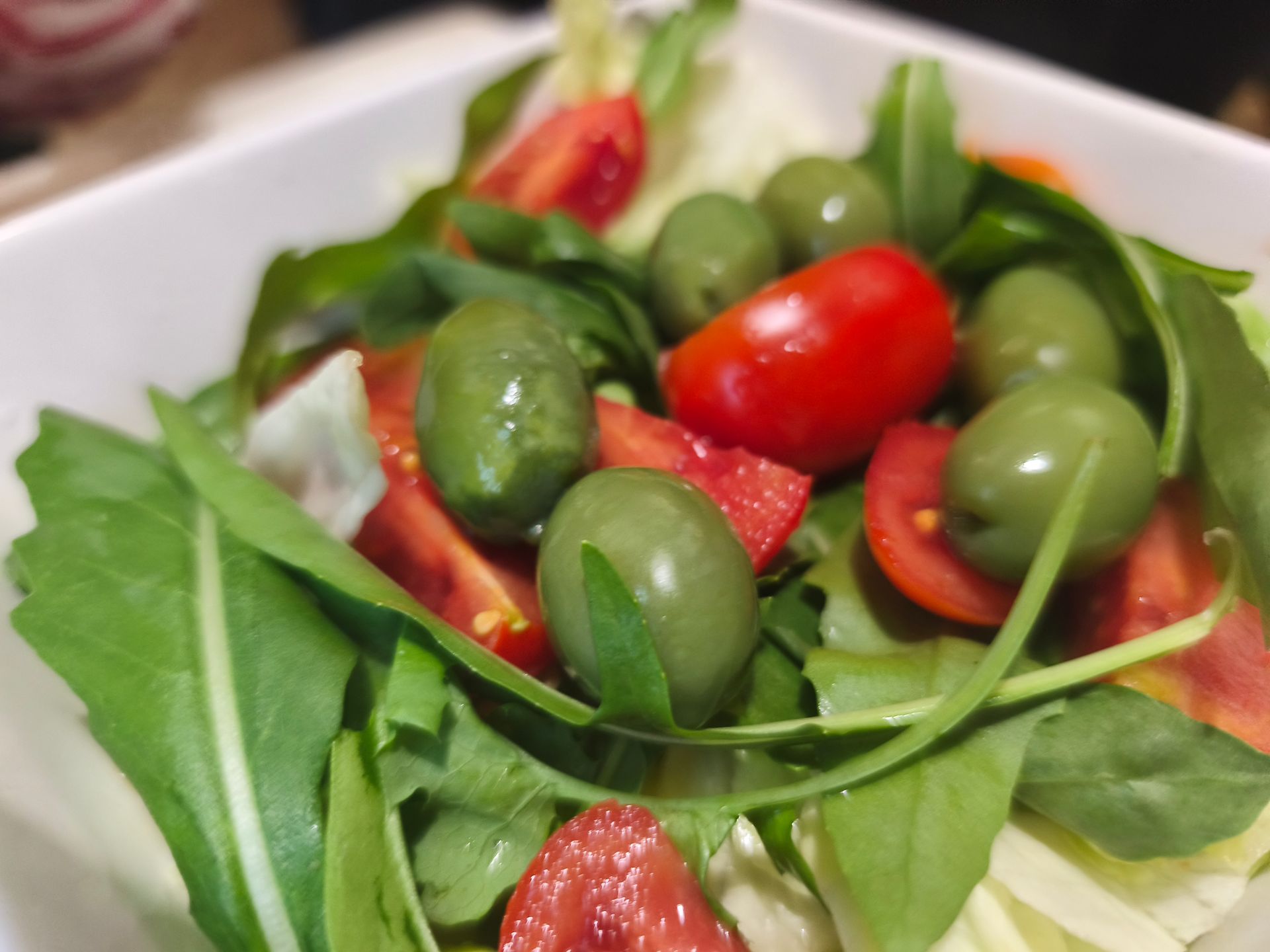 Primo piano di un'insalata fresca con foglie verdi, olive e pomodorini in una ciotola bianca.