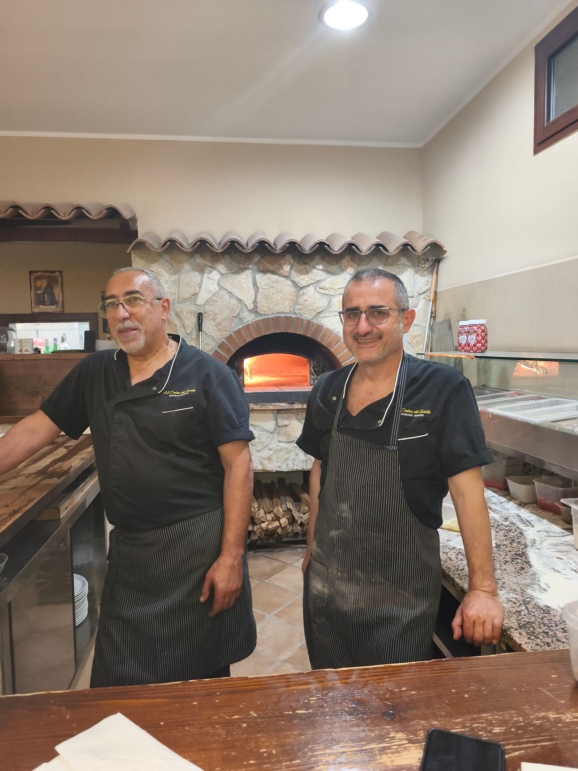 Due uomini con grembiuli neri posano accanto a un forno a legna in una cucina rustica.