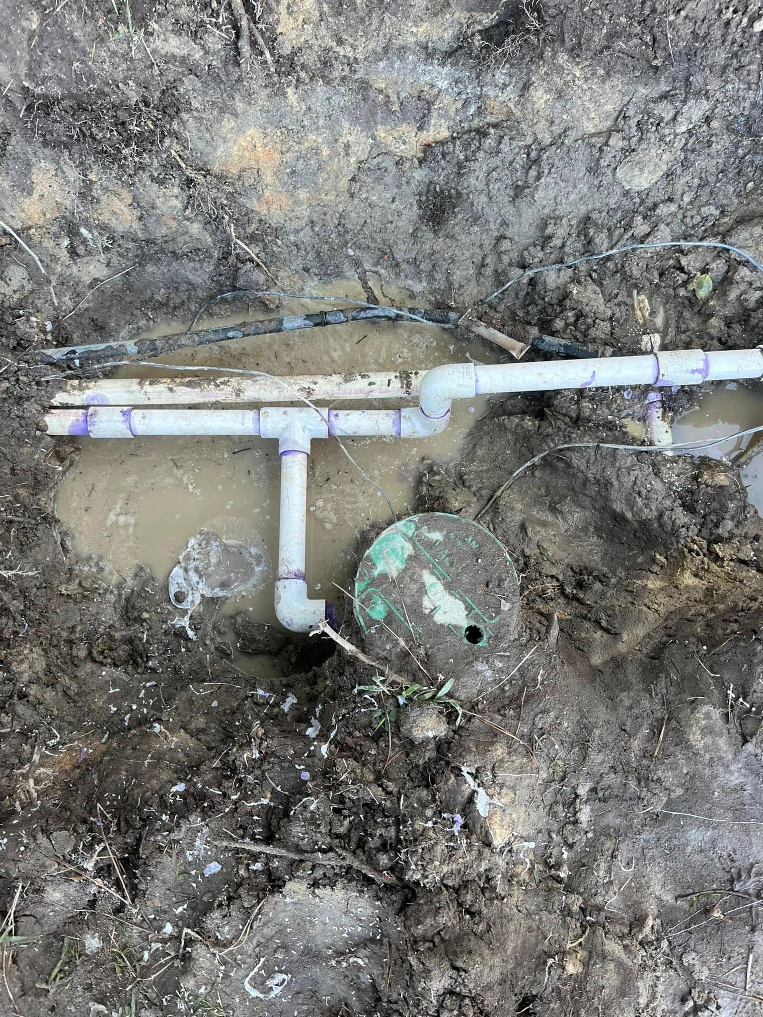 White PVC pipe with a T-joint and valve in muddy soil beside a dirt wall