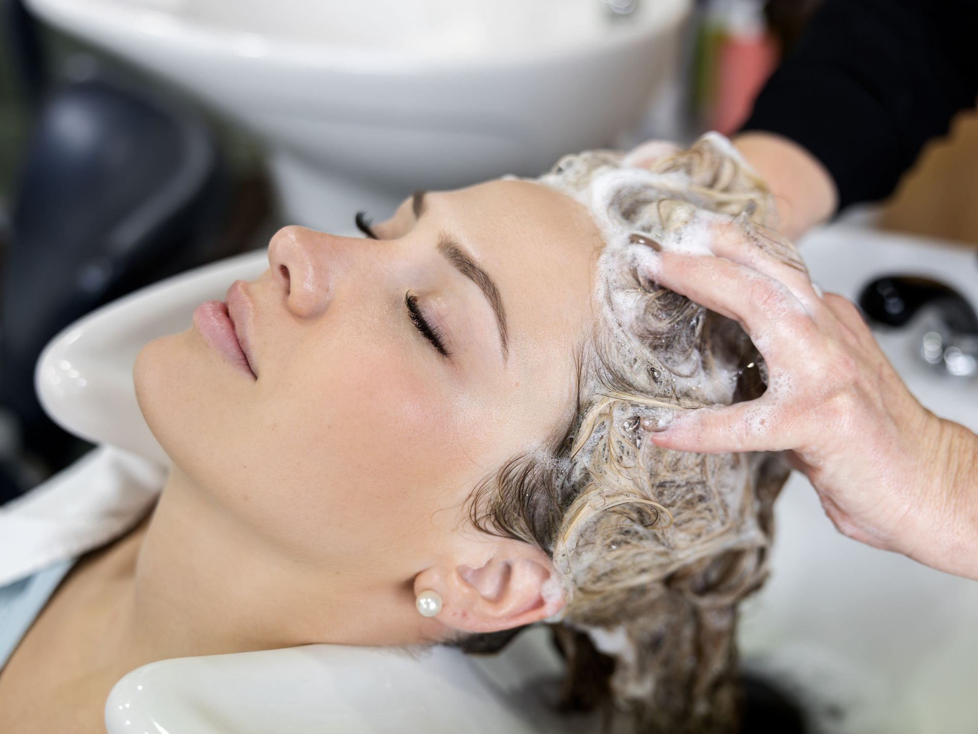 A person with eyes closed leans back in a salon sink as hands massage soapy lather into their hair. A person with eyes closed leans back in a salon sink as hands massage soapy lather into their hair.
