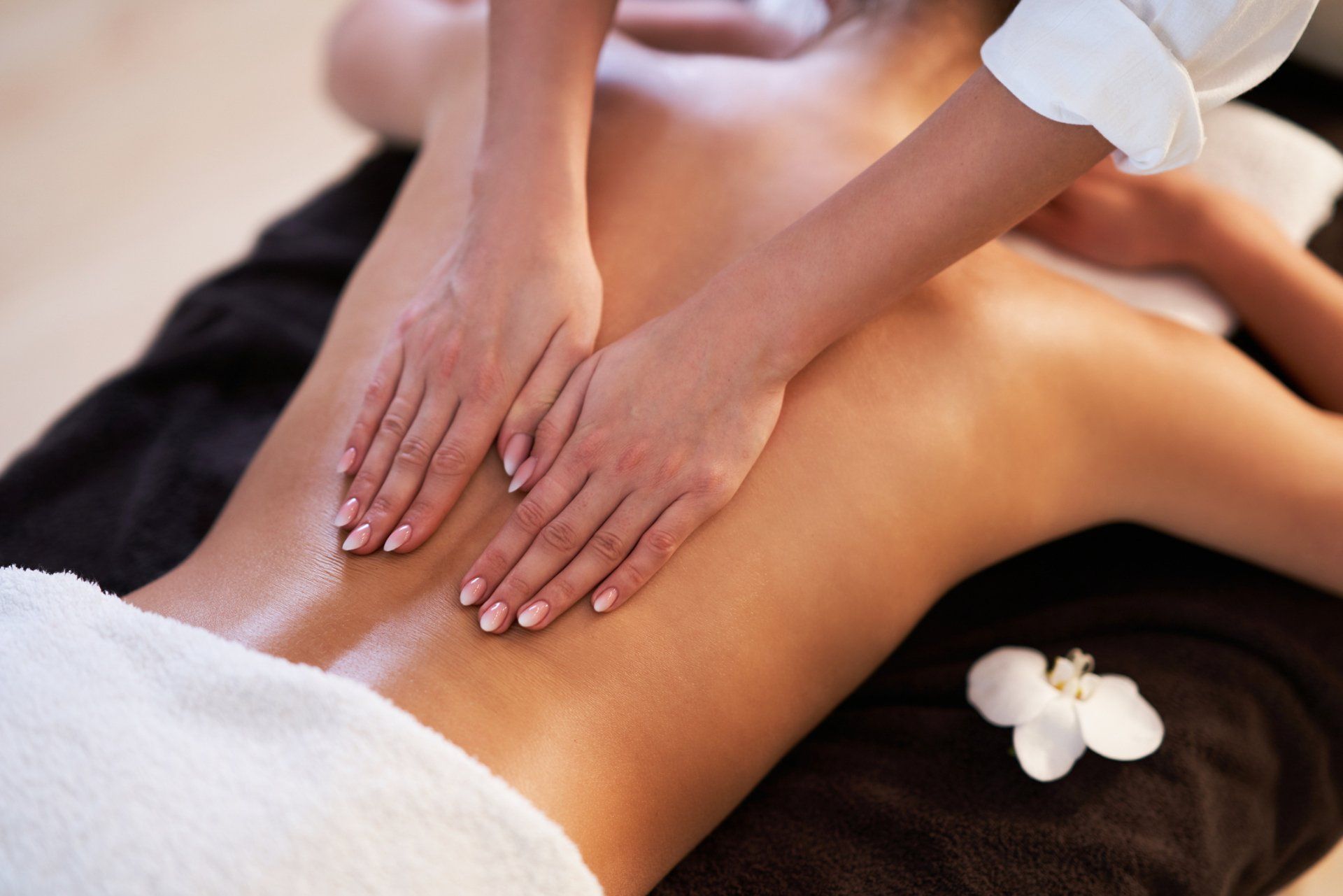 Hands performing a massage on a person's oiled, bare back with a white flower on a black towel nearby. Hands performing a massage on a person's oiled, bare back with a white flower on a black towel nearby.