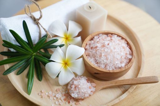 A wooden tray holding pink Himalayan salt in a bowl and spoon, white plumeria flowers, a towel, and a candle. A wooden tray holding pink Himalayan salt in a bowl and spoon, white plumeria flowers, a towel, and a candle.