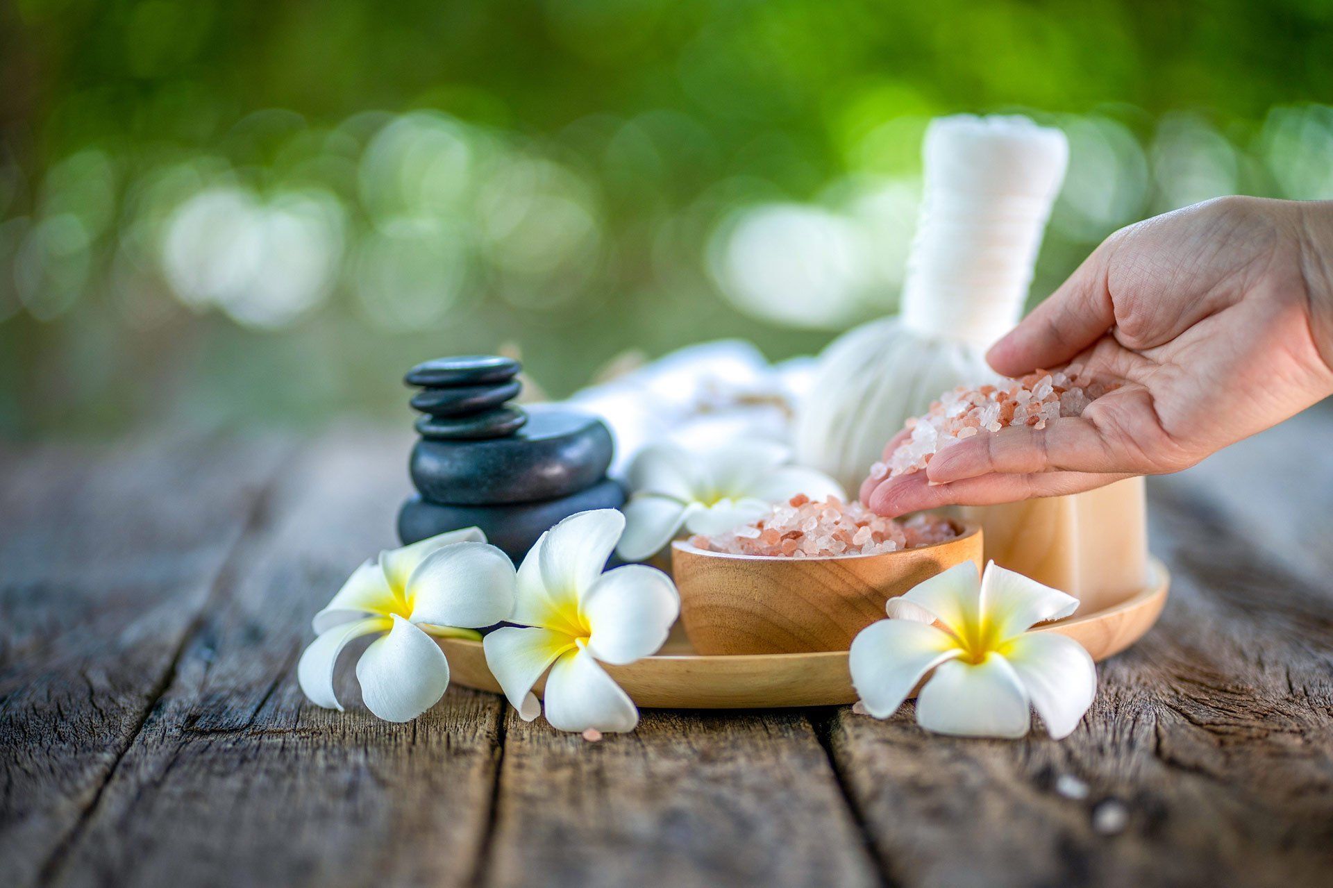 A hand holds pink Himalayan salt near spa elements: a stack of stones, plumeria flowers, and a herbal compress on wood. A hand holds pink Himalayan salt near spa elements: a stack of stones, plumeria flowers, and a herbal compress on wood.