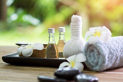Spa items on a wooden tray: two glass bottles of oil, a white massage compress, a rolled towel, and frangipani flowers. Spa items on a wooden tray: two glass bottles of oil, a white massage compress, a rolled towel, and frangipani flowers.