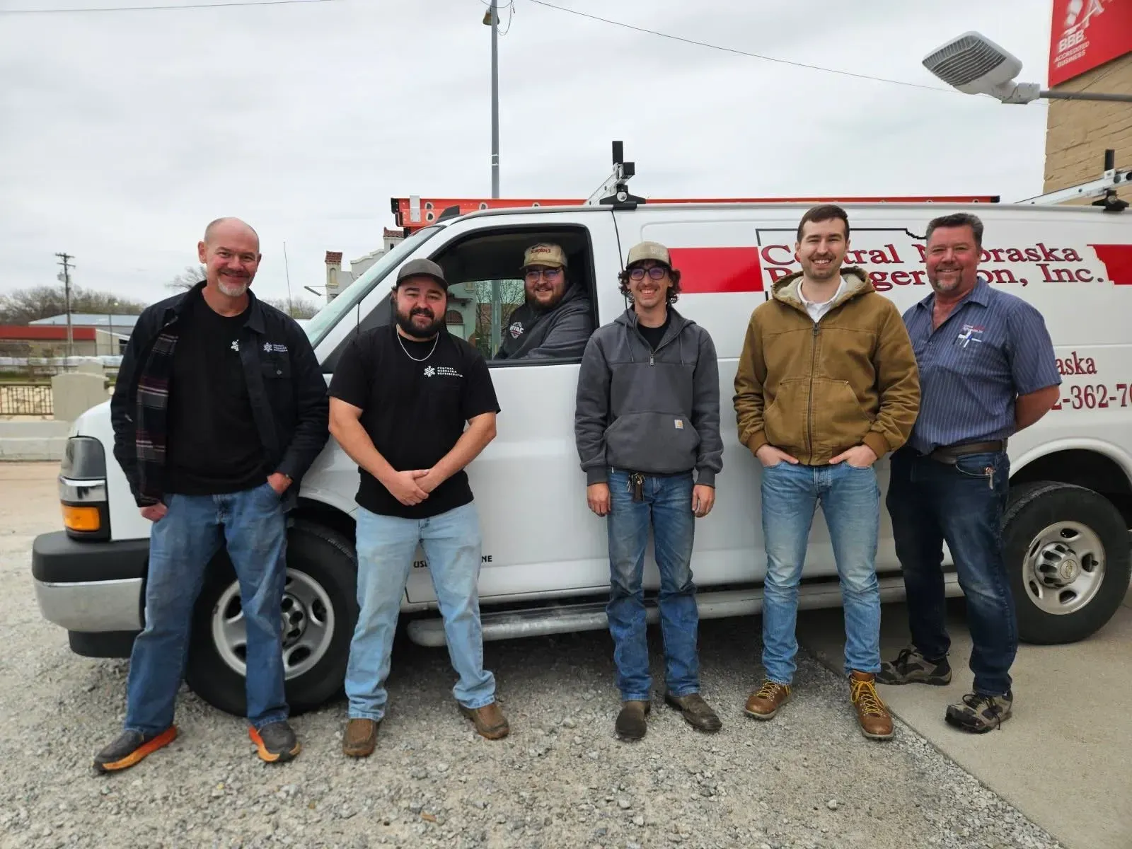 Five men standing in front of a white work van with roof equipment, outdoors on a cloudy day
