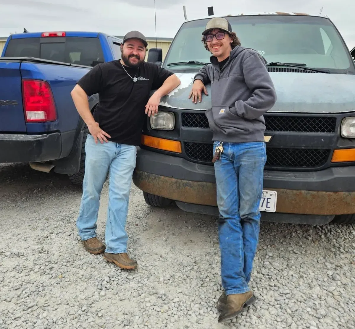 Two people leaning on vehicles in a gravel lot, one black truck and one blue truck behind them.