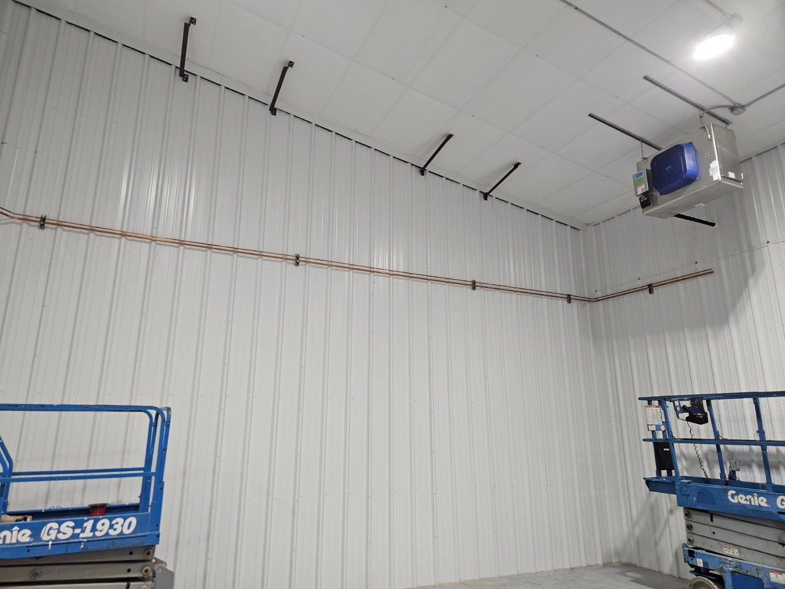 White industrial room with high ceiling, wall-mounted wiring, and blue equipment lifts in the corners