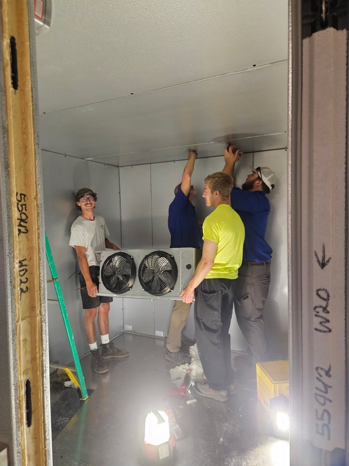 Workers installing a ceiling in an unfinished room with two fans and tools on the floor.