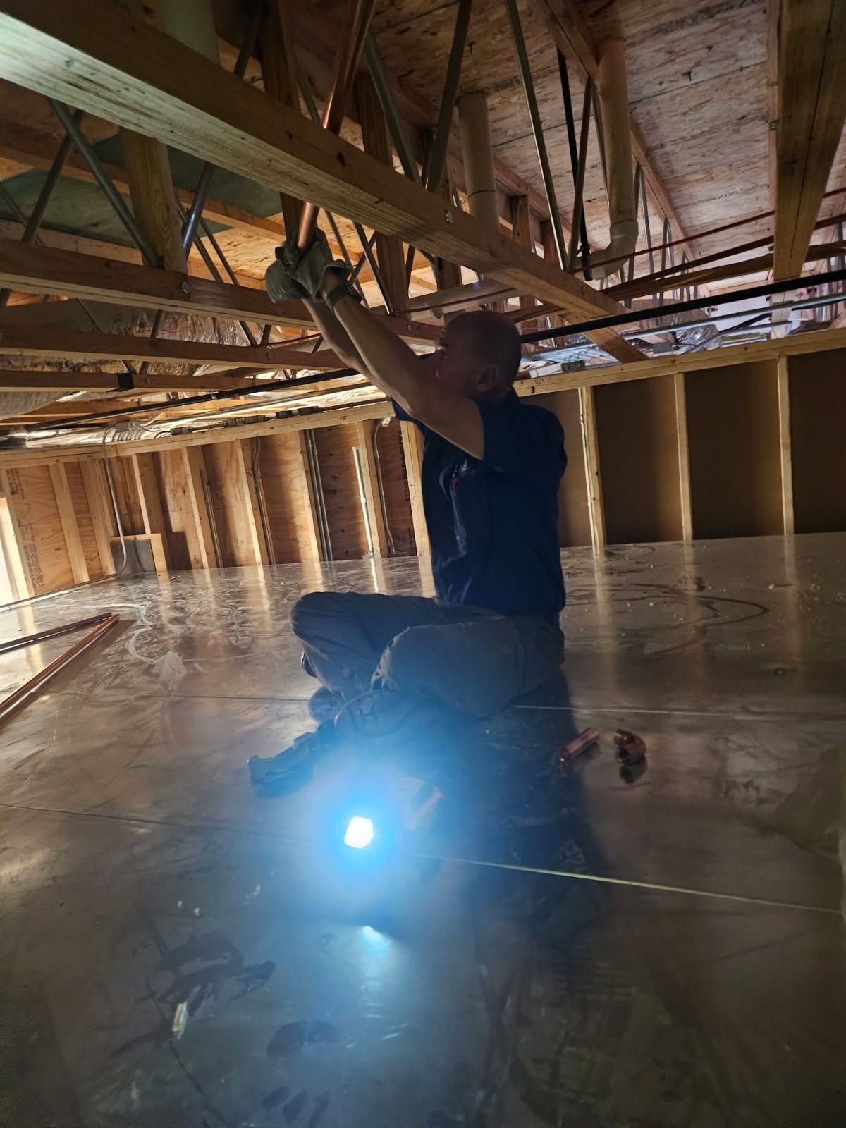 Worker kneeling in an unfinished attic, inspecting exposed ceiling framing with a bright flashlight