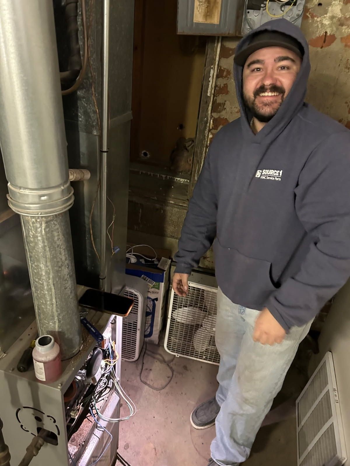 Smiling worker in a gray hoodie stands in a narrow utility room beside pipes and HVAC equipment.
