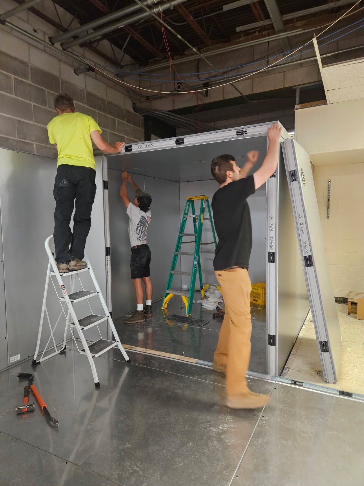Three people assemble a large metal frame inside an industrial room, using ladders and tools.